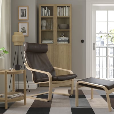 Chaise POÄNG en cuir marron, structure en bois, dossier haut. À côté de l’étagère en bois, près de la fenêtre. Confortable et spacieux.