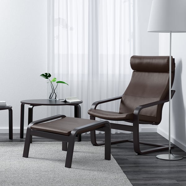 Salon moderne avec chaise POÄNG en cuir marron, repose-pieds, table d’appoint, plante, livre, lampe et tapis.