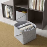 Deux cubes de rangement en tissu gris, l’un contenant des boîtes, l’autre du fil, à côté d’une étagère grise avec des livres.