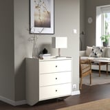 Commode blanche à 3 tiroirs RACKNÄS dans un coin avec lampe, vase, art ; près du canapé, de la table, de la bascule.