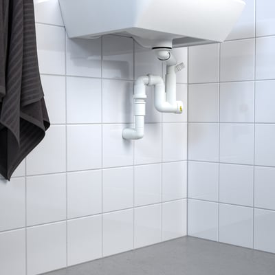 Évier blanc avec siphon d’eau RÄNNILEN visible et tuyauterie flexible contre le mur carrelé.
