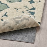 Un tapis multicolore à poils longs avec un motif 3D texturé. En dessous, une sous-couche de tapis antidérapante est visible, ce qui améliore la sécurité et le confort.