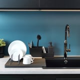 Évier de cuisine moderne avec un séchoir à vaisselle contenant des assiettes et des tasses blanches. Un distributeur de savon et une brosse à récurer se trouvent à proximité, contre un dosseret bleu.