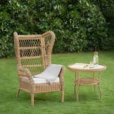Chaise en osier et table d’appoint sur pelouse, rotin, coussin, couverture, verrerie, livre, buissons verts.