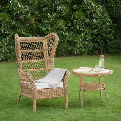 Chaise en osier et table d’appoint sur pelouse, rotin, coussin, couverture, verrerie, livre, buissons verts.