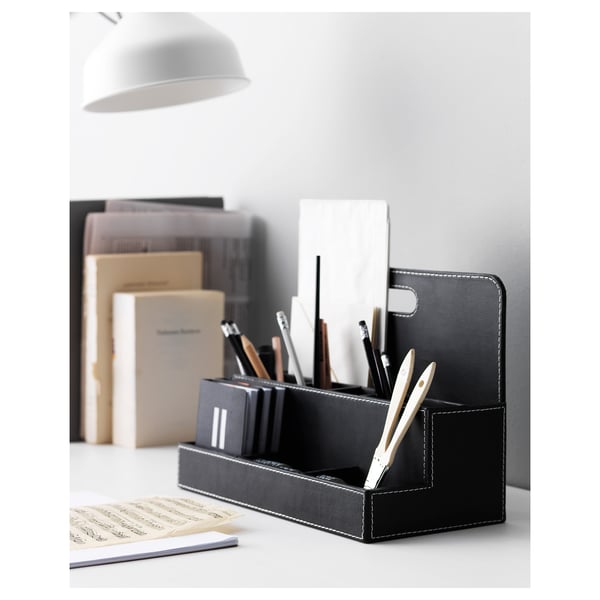 Organisateur de bureau noir avec compartiments pour stylos, marqueurs et règle. Fabriqué en carton laminé. Entouré de livres, de lampes et de papiers.