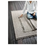 Personne disposant un tapis en coton RÖRKÄR beige et noir sur un plancher en bois foncé.