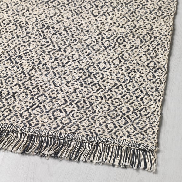 Tapis texturé, tissé en noir et blanc avec des bords à franges, exposé sur une surface plane.