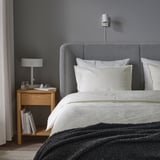 Chambre moderne avec tête de lit grise, literie blanche, couverture sombre, table de chevet en bois, lampe, livres et applique murale.
