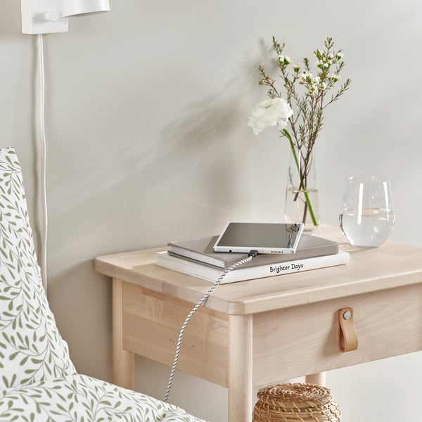 Table de chevet avec un ordinateur portable, des livres, des fleurs et un câble de charge.