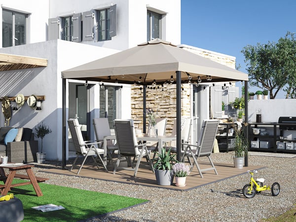 Patio arrière avec terrasse RUNNEN beige, chaises grises, table et auvent avec lumières, entouré de gravier et de verdure.
