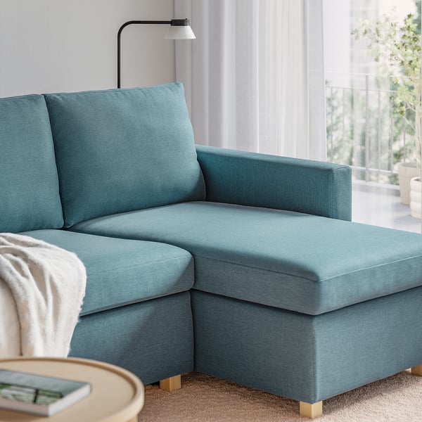 Canapé-lit bleu avec accoudoirs, coussins, et une table basse en bois avec un livre et une couverture à proximité.