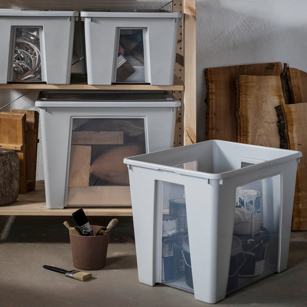 La boîte de rangement SAMLA blanche avec fenêtres transparentes contient des boîtes de peinture, placées au sol près d’étagères en bois contenant d’autres boîtes SAMLA , du bois et des outils.