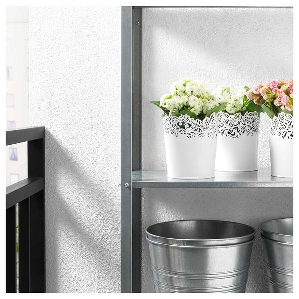 Trois pots décoratifs blancs avec des fleurs sur une étagère en métal. Deux grands seaux métalliques en dessous.