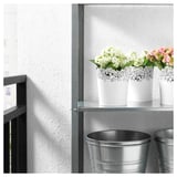 Trois pots décoratifs blancs avec des fleurs sur une étagère en métal. Deux grands seaux métalliques en dessous.