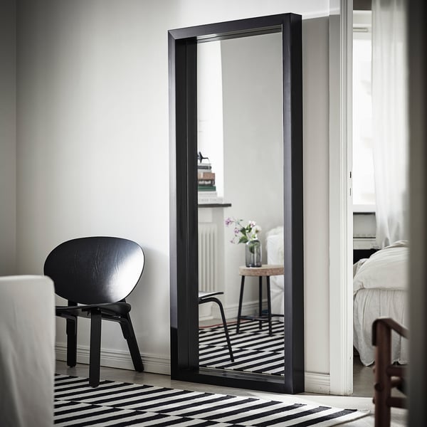 Grand miroir à cadre noir appuyé contre le mur reflétant la pièce minimaliste avec chaise noire et tapis rayé.