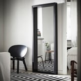 Grand miroir à cadre noir appuyé contre le mur reflétant la pièce minimaliste avec chaise noire et tapis rayé.