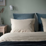 Chambre confortable avec coussin beige, oreillers bleus, mur vert, lampe, livres, vase.