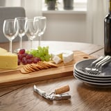 Table en bois avec du fromage, des raisins, des craquelins, des verres à vin, des assiettes, des fourchettes, un tire-bouchon et une bouteille, prête pour un repas.