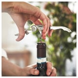 Une personne utilisant un tire-bouchon SEGELFISK en argent pour ouvrir une bouteille de vin, mettant en valeur son design épuré et sa praticité.