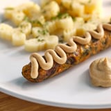 Une assiette blanche contient des saucisses brunes arrosées d’une trempette crémeuse, à côté d’une salade de pommes de terre dorées.