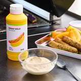 Imaginez une bouteille de moutarde jaune étiquetée SENAP MILD à côté d’un bol de sauce et d’un plateau de saucisses sur un gril.