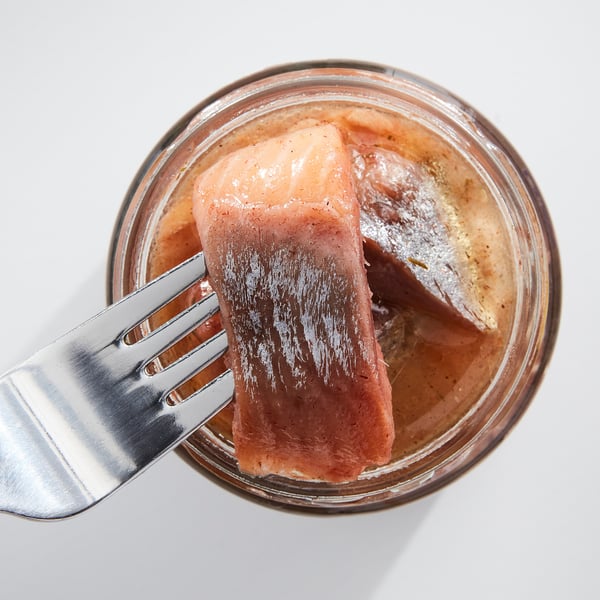 Un pot de hareng mariné rose clair, tenu par une fourchette. Récipient en verre transparent, fourchette en argent.