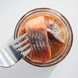 Un pot de hareng mariné rose clair, tenu par une fourchette. Récipient en verre transparent, fourchette en argent.