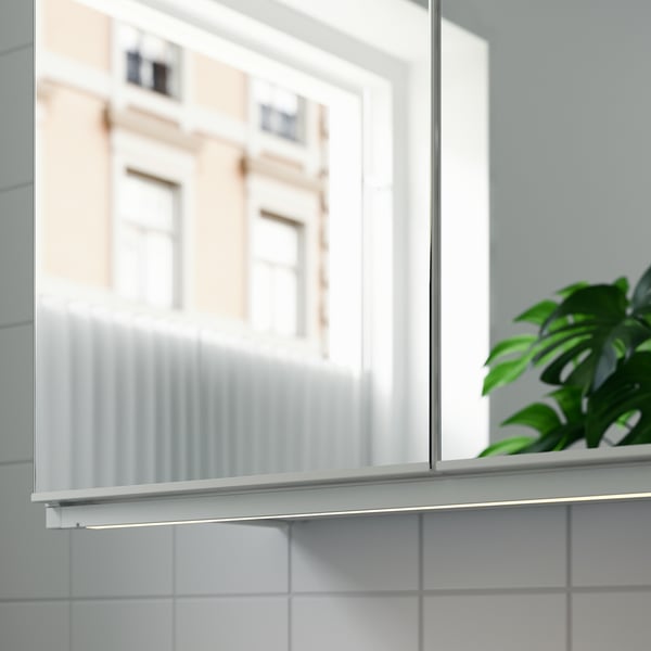 Armoire blanche avec lumière led, bâtiment réfléchissant, plante verte sur carreaux.