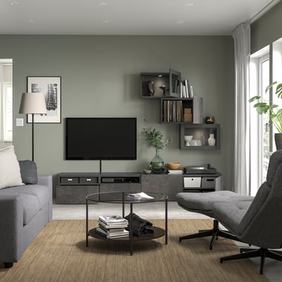 Salon moderne avec meuble TV SINDVIK gris, canapé gris, table basse en verre et fauteuil gris. Mur d’accent vert avec étagères.