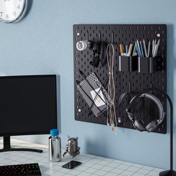 Panneau perforé mural noir avec crochets pour tenir des écouteurs, des stylos et des câbles. En dessous, un bureau avec un moniteur, un clavier et d’autres objets.