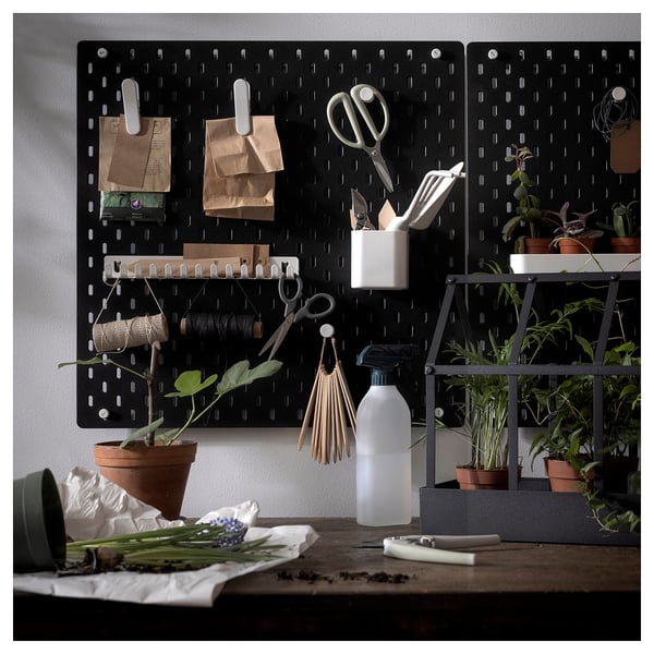 Panneau perforé SKÅDIS noir avec divers ustensiles de cuisine et plantes attachés, y compris des ciseaux, des sacs et un vaporisateur, disposés contre un mur.