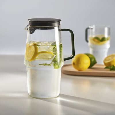 Pichet en verre transparent avec couvercle et anse noirs, rempli de tranches de citron et de glaçons. Idéal pour servir des boissons fraîches.