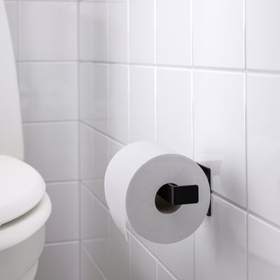 Porte-papier toilette noir monté sur un mur carrelé blanc, tenant un rouleau de papier toilette blanc, près de toilettes blanches.