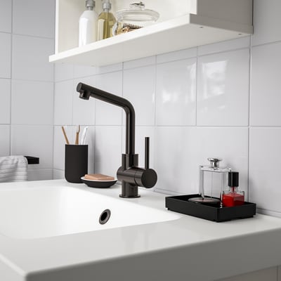 Un petit organisateur de salle de bain SKOGSVIKEN noir se trouve à côté du lavabo, contenant les articles de toilette et le maquillage.