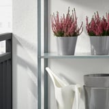 Deux fleurs roses en pot sur une étagère en métal à côté d’un arrosoir blanc.