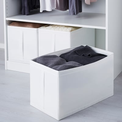 Des boîtes de rangement blanches dans un placard, l’une remplie de vêtements gris foncé pliés, soigneusement empilées pour un accès organisé à la garde-robe.