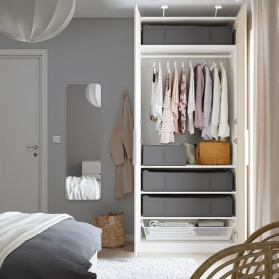Chambre avec des caisses de rangement SKUBB blanches dans une armoire ouverte, contenant des vêtements pliés et un sac. Un miroir et un panier en osier se trouvent à côté de l’armoire.