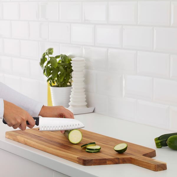Une personne tranche du concombre sur une planche à découper en bois sur un comptoir de cuisine blanc.