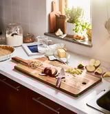 Une planche à découper en bois avec un couteau et des tranches de pomme se trouve sur un comptoir de cuisine blanc, entourée d’ingrédients de cuisson.