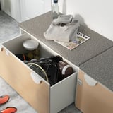 Banc de rangement ouvert, beige et gris, panneaux imitation bois, équipement de sport blanc à l’intérieur, sweat-shirt gris sur le dessus.