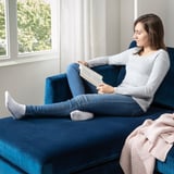 Une femme lit sur un canapé SMEDSTORP en velours bleu près de la fenêtre, les pieds en l’air, profitant du confort d’une couverture douillette à proximité.