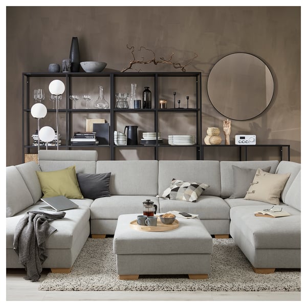 Salon moderne avec canapé sectionnel beige, oreillers gris, table basse en bois, étagère noire avec décor, miroir rond et tapis de couleur claire.