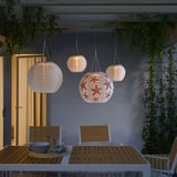 Trois lampes SOLVINDEN avec abat-jour en étoile de mer brillent au-dessus d’une table en bois, créant ainsi une configuration de salle à manger en plein air confortable et écologique.
