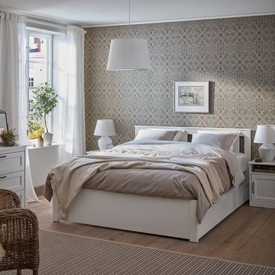 Chambre propre avec SONGES et lit blanc, papier peint à motifs, draps gris, tables de chevet et une chaise en osier.