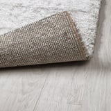 Un tapis gris enroulé avec des poils épais et texturés, posé sur un parquet en bois clair. Le tissage dense suggère le confort et l’absorption acoustique.