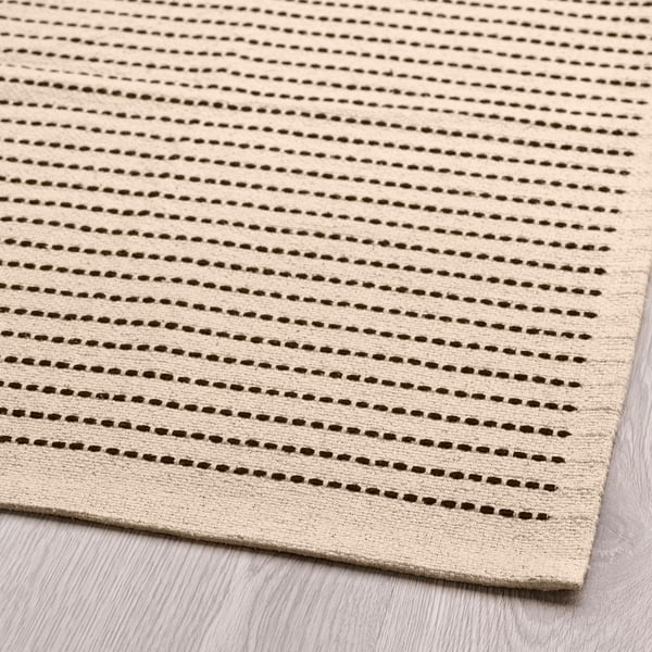 Tapis tissé à plat, ton naturel avec des rayures sombres, posé sur un plancher en bois. Coton de haute qualité, design durable et propre.