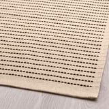 Tapis tissé à plat, ton naturel avec des rayures sombres, posé sur un plancher en bois. Coton de haute qualité, design durable et propre.