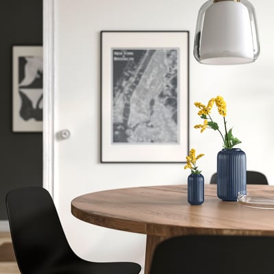Salle à manger moderne avec table en bois, chaises noires, deux vases bleus avec des fleurs jaunes, affiche en noir et blanc, lampe suspendue.