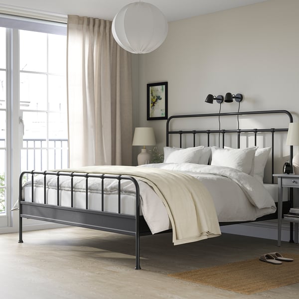 Lit STJÄRNÖ noir avec tête de lit haute, literie blanche/beige, grande fenêtre avec rideaux beiges, tapis gris, 3 appliques murales.